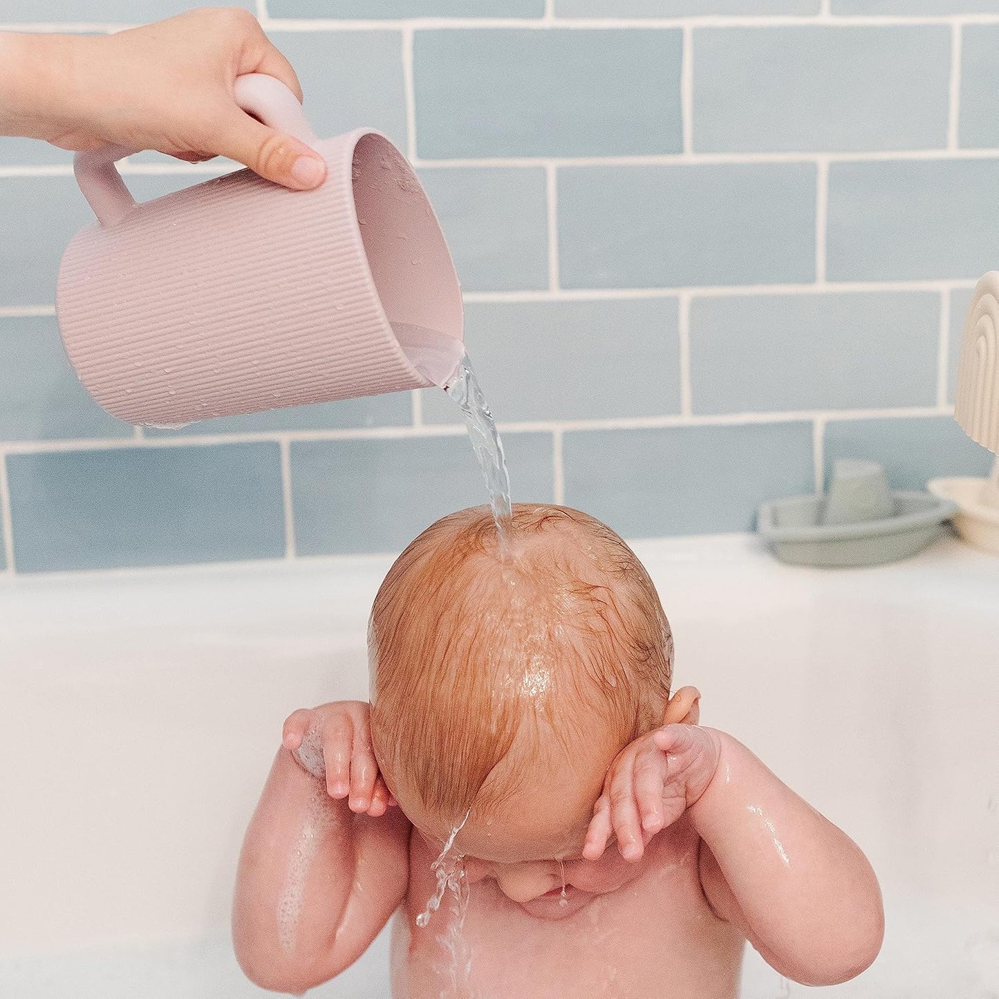 Baby being bathed with water from a pink cup against a tiled bathroom wall.