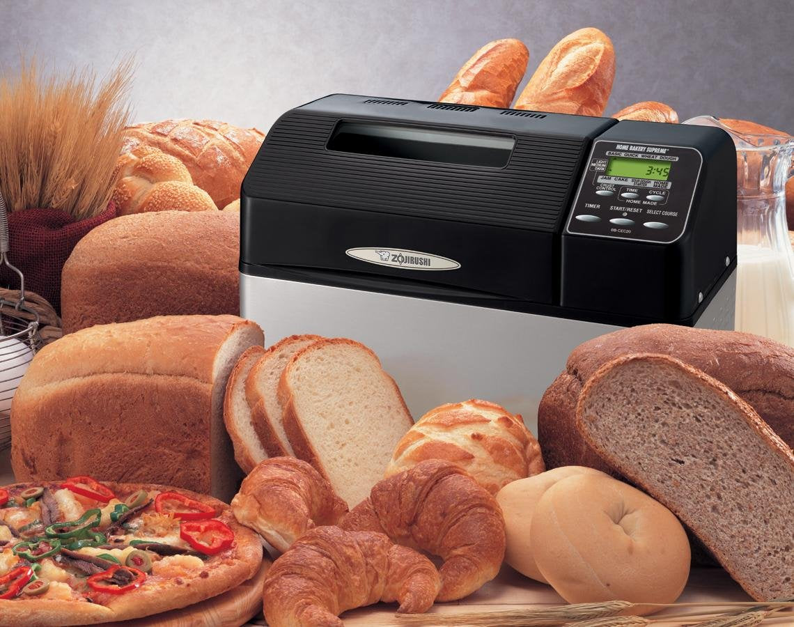 Zojirushi Home Bakery Supreme surrounded by various types of bread and a pizza on a wooden surface.