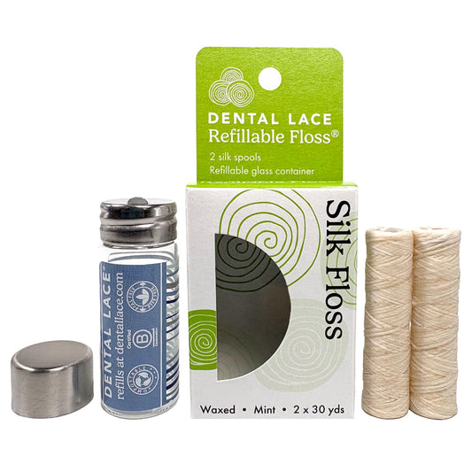 Dental lace refillable floss set including a glass bottle with a blue label and silver cap, a green and white hanging card, a white box labeled Silk Floss, and two beige silk thread spools on a white background.
