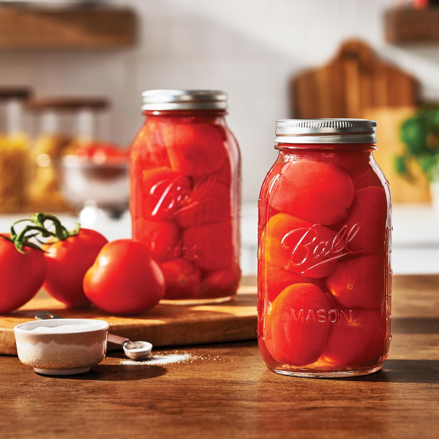 Two jars of canned tomatoes on a wooden table with fresh tomatoes and kitchen items in the background.