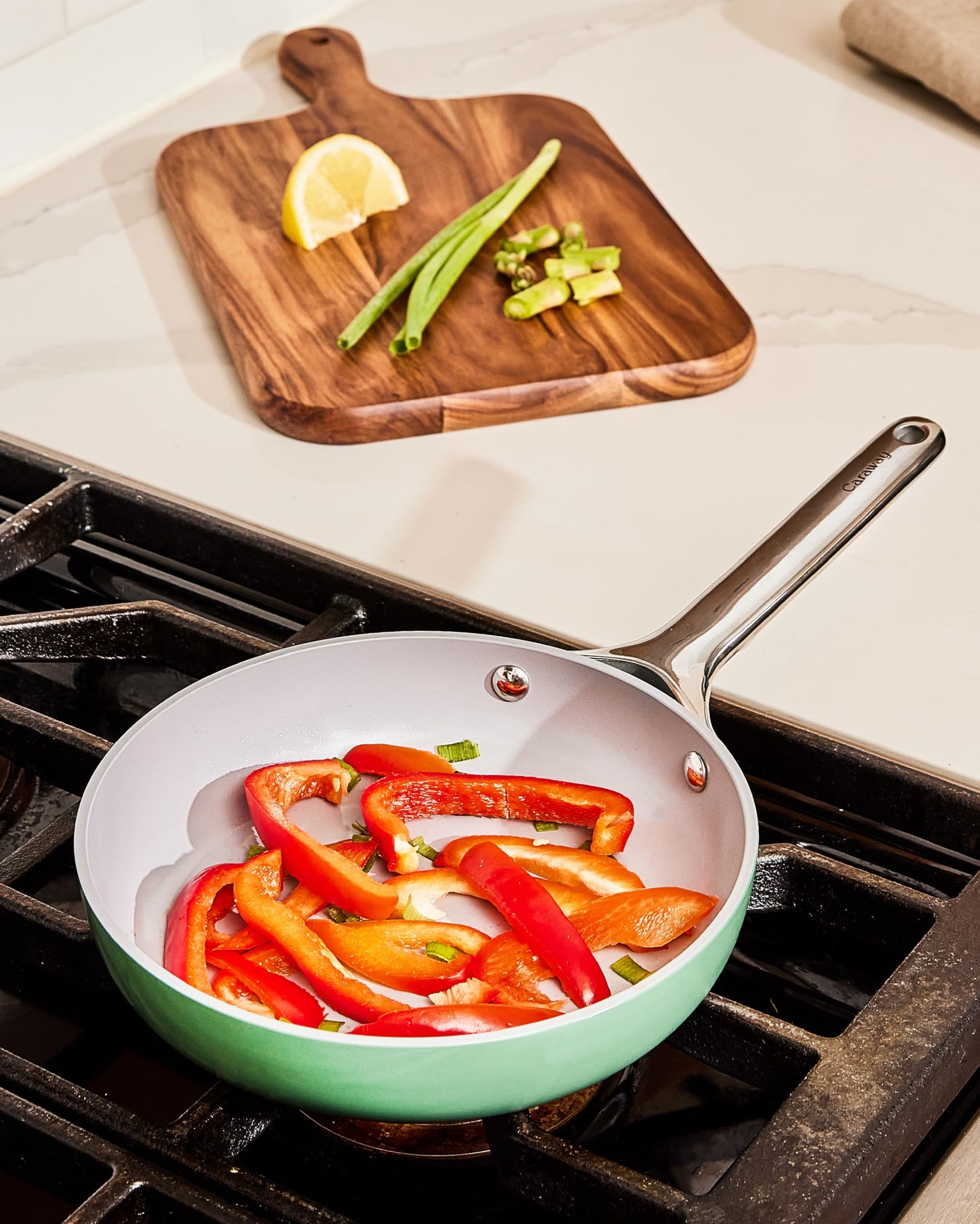 Frying pan with sliced red peppers on a stovetop, wooden cutting board with lemon and green onions in the background.