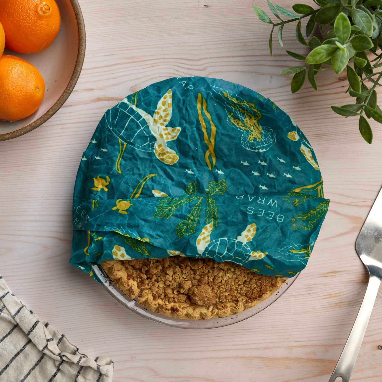 Pie wrapped in a reusable beeswax wrap on a wooden table with oranges and a fork.