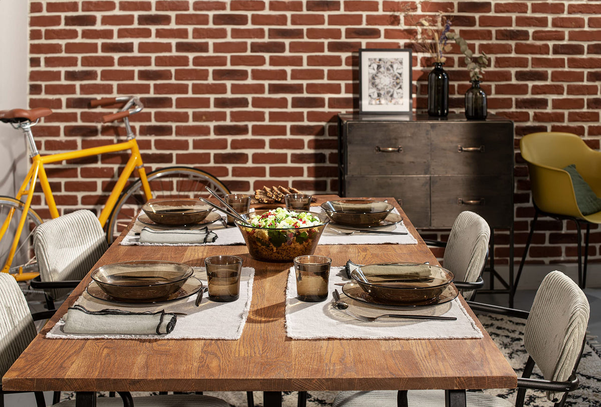 Dining table set with Duralex Lys Clear Glass Dinnerware Set, 18-Piece (Service for 6) against a brick wall with a bicycle and furniture in the background.