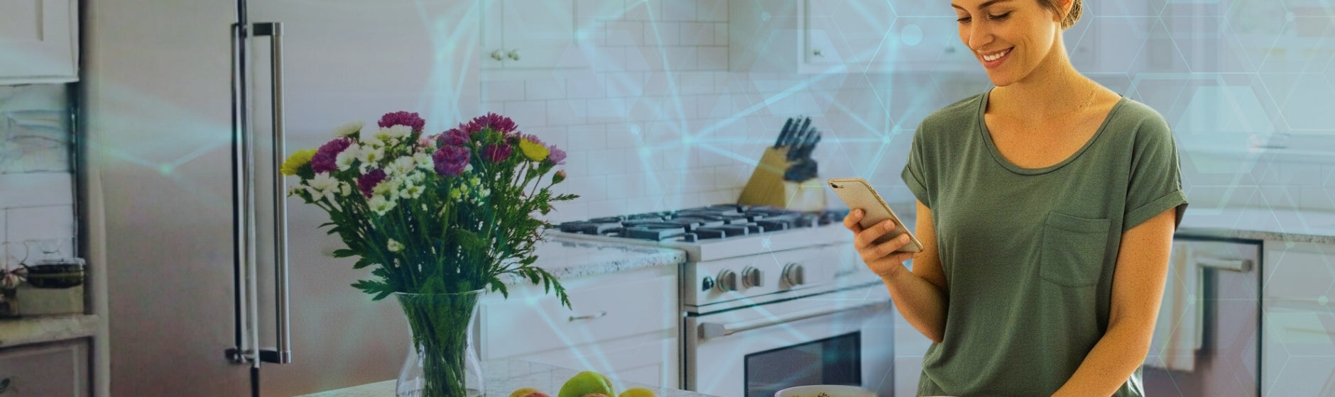 Woman using a smartphone in a kitchen with a vase of flowers and fruits on the counter.