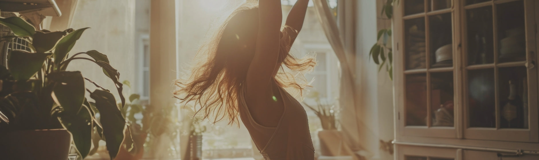 Person with long hair standing in a sunlit room with plants and furniture.