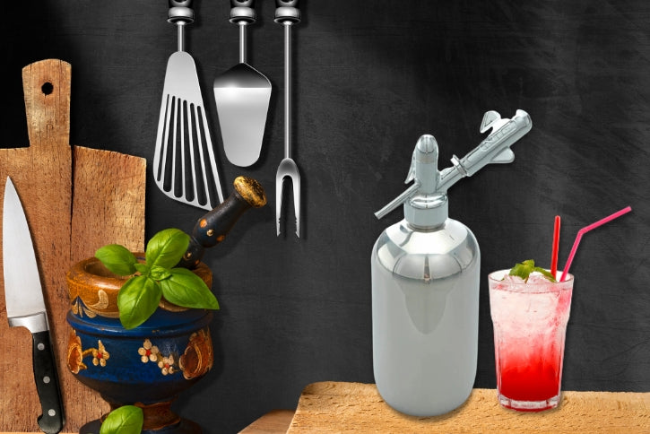 A stylized culinary scene set against a dark, chalkboard-like background. On the left, a wooden cutting board stands upright next to a chef's knife and a decorative blue and gold mortar and pestle holding fresh basil leaves. Three stainless steel kitchen utensils hang above them. On the right, resting on a wooden surface, is a shiny stainless steel soda siphon next to a tall glass filled with ice, a bright red beverage, a sprig of mint, and two red straws.